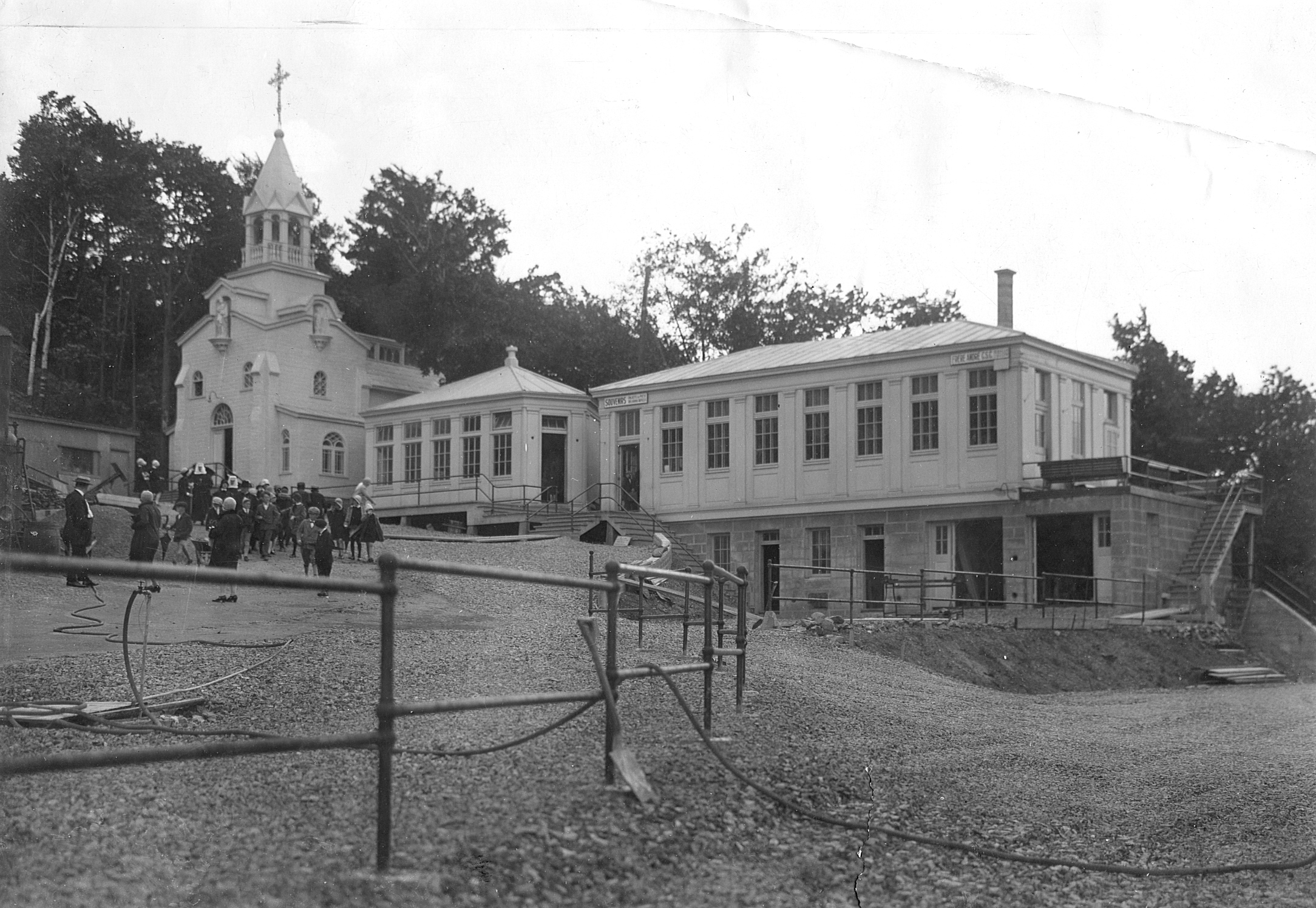 Kiosque des plans et magasin d’objets de piété en 1927. En 1953-1954, ces deux bâtiments sont réunis et agrandis, donnant l’actuel pavillon des pèlerins. La chapelle est déménagée en 1954. Photographie en noir et blanc.